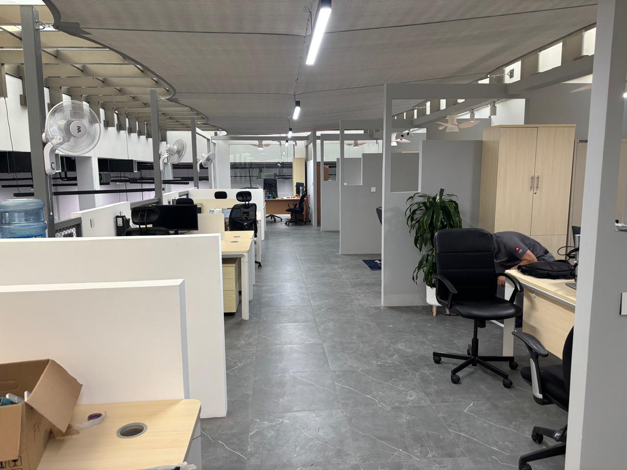 Renovated warehouse office with cubicles, wood desks, grey flooring, ceiling lights, and view into the warehouse